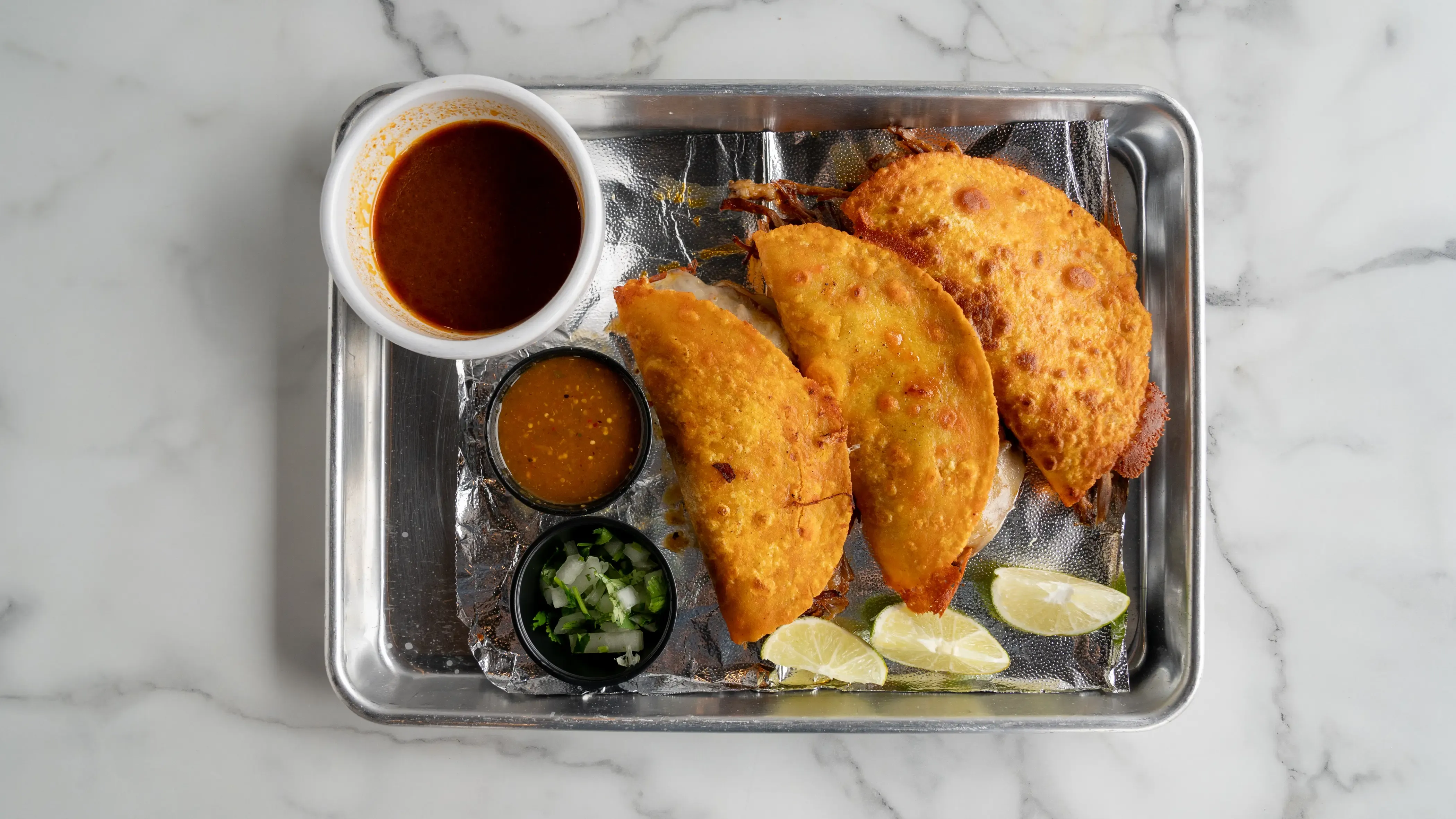 Three corn tortillas filled with slow-cooked, spiced beef birria. Served with rich consommé broth for dipping, fresh cilantro, diced onions, lime wedges, and spicy sauce